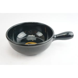 May include: A black ceramic bowl with a handle. The bowl has a glossy finish and a round shape. The handle is attached to the side of the bowl. The bowl is sitting on a white surface.