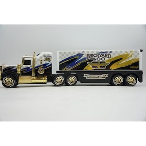 May include: A Buddy L toy semi-truck featuring a white cab and trailer with blue and gold graphic accents. The side displays the text Brickyard 400, Inaugural Race August 6, 1994, and a checkered flag pattern. The truck has gold-colored wheels and trim.