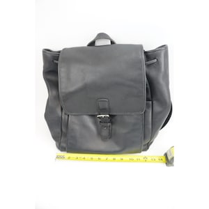 May include: A black leather backpack with a top flap, a central buckle strap, and side pockets. A yellow measuring tape is positioned along the bottom, showing a width of approximately 14 inches for the base of the bag.