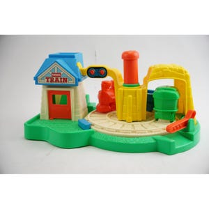 May include: A colorful Playskool Junior Train set. The set includes a train station with a blue roof, a yellow crane, a green train car, and a red train. The base is green with a circular track.