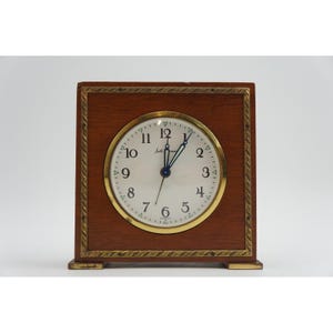 May include: A square wooden mantel clock with a warm brown finish and a gold-toned metal border featuring a rope-like texture. The white clock face displays black Arabic numerals, blue hands, and the brand name Seth Thomas in black script lettering.