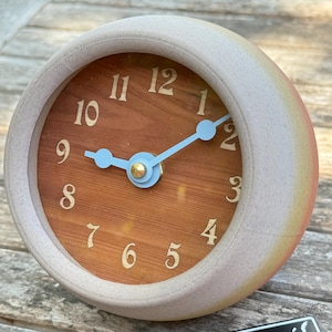 May include: A round, cream-colored analog clock with a wood-grain face. The clock has light blue hands and gold accents. The numbers are in a serif font. The clock sits on a wooden surface. A small black and white sign with the text "WavyTail" is in the foreground.