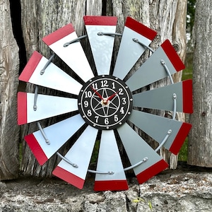 May include: A decorative windmill clock with a gray and red color scheme. The clock face is black with white numbers and hands. The windmill blades are gray with red tips. The clock is mounted on a weathered wooden fence.