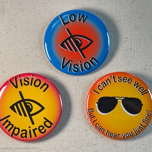 Peut inclure: Trois badges ronds avec une finition brillante. Le badge du haut est bleu et rouge avec une icône d'œil barré et le texte Low Vision. Celui en bas à gauche est jaune et rouge avec un œil barré et Vision Impaired. Celui en bas à droite est jaune et orange avec des lunettes de soleil et le texte I can't see well But I can hear you just fine!