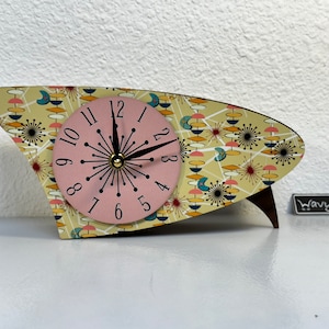 May include: A retro-style clock with a light pink face and black hands. The clock's body has a unique, asymmetrical shape and is decorated with a colorful mid-century modern pattern. The clock is supported by a dark brown wooden base.