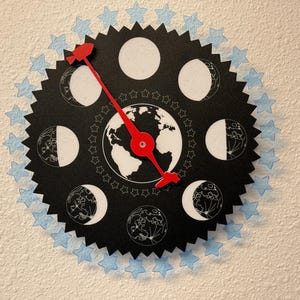 May include: A unique wall clock featuring a black circular design with a gear-like edge. The clock face displays the phases of the moon, a globe, and red hands. Light blue star accents surround the clock, creating a celestial theme.