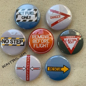 May include: Set of seven round buttons with various aviation safety warnings. The buttons are white, gray, blue, and red with black, yellow, and white text. The text on the buttons includes "Jet Fuel Only", "Danger Jet Intake", "No Step", "Remove Before Flight", "Danger Ejection Seat", "Danger Propeller", and "Rescue".