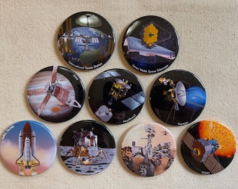 May include: Nine round, black and white, space-themed buttons with images of spacecraft and planets. The buttons feature the names of the spacecraft, including Space Shuttle, International Space Station, Juno, Hayabusa2, Voyager, James Webb Space Telescope, Apollo 11 Lunar Lander, Curiosity Rover, and SOHO.