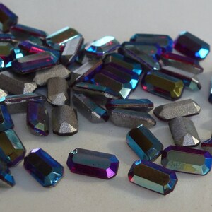 Op de afbeelding: Een verzameling van iriserende blauwe en paarse glazen edelstenen in een rechthoekige vorm. De edelstenen hebben een facetgeslepen ontwerp en zijn verspreid over een wit oppervlak.