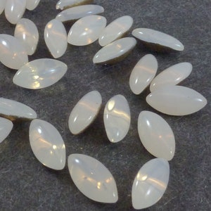 Può includere: Una collezione di cabochon di vetro bianco ovali e iridescenti. I cabochon sono sparsi su una superficie grigia.