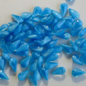 May include: A large group of light blue teardrop-shaped glass beads. The beads are scattered on a white surface.