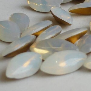 Può includere: Un mucchio di strass ovali in vetro bianco con retro in foglia d'oro. Gli strass sono sparsi su una superficie bianca.