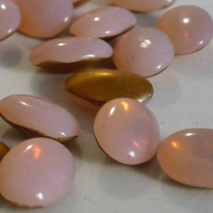 Può includere: Una collezione di bottoni ovali di colore rosa pallido con una finitura perlescente. Alcuni dei bottoni hanno un retro di colore dorato.