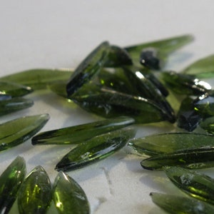 May include: A collection of olive-green, leaf-shaped glass beads. The beads are elongated with pointed ends and a faceted surface, reflecting light. These decorative beads are suitable for jewelry making and craft projects.