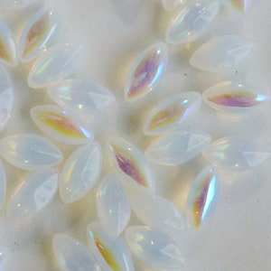Può includere: Una collezione di perline di vetro bianco iridescente di forma ovale. Le perline hanno una finitura madreperlacea e sono sparse su una superficie bianca.