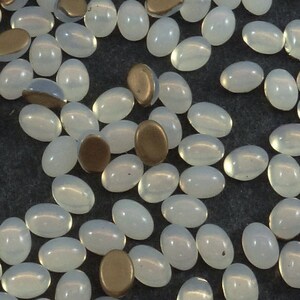Peut inclure: Un ensemble de perles ovales opalescentes et de perles ovales de couleur bronze, dispersées sur une surface sombre. Ces perles sont probablement destinées à la fabrication de bijoux ou à des projets d'artisanat. Les perles mesurent environ 1 cm de long.
