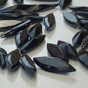 Peut inclure: Un ensemble de perles en verre noires facettées en forme de larme. Les perles sont disposées de manière aléatoire sur une surface blanche.
