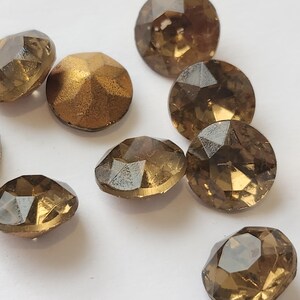May include: A selection of faceted, round, amber-coloured rhinestones. The gemstones have a metallic sheen and are scattered on a white surface. The rhinestones vary slightly in size and shape, with multiple facets.