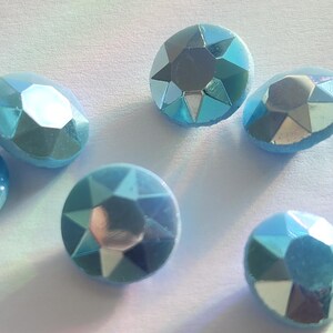 May include: Close-up of several light blue, faceted gemstones. The gems have a reflective, iridescent quality, with facets catching the light. The shapes vary from round to oval, suggesting they could be used for jewellery or craft projects.