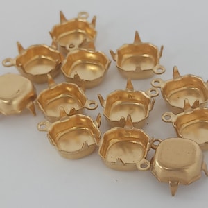 Op de afbeelding: Een groep van 12 goudkleurige metalen instellingen voor cabochons. Elke instelling heeft vier pinnen en een lus om een ​​ketting of springring aan te bevestigen.