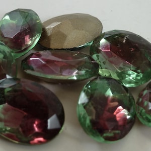 Puede incluir: Un grupo de ocho cabochones de vidrio iridiscente de forma ovalada. Los cabochons son una mezcla de verde y morado.