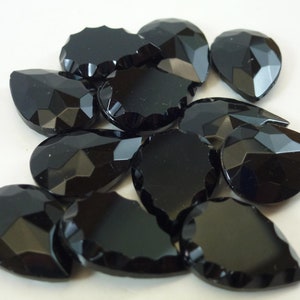 Puede incluir: Un grupo de piedras de cristal negras en forma de lágrima. Las piedras tienen un diseño facetado y están esparcidas sobre una superficie blanca.
