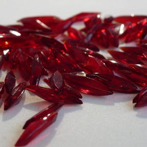 May include: A large quantity of red, faceted, glass teardrop-shaped beads. The beads are scattered on a white surface.