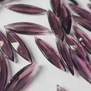 May include: Close-up of numerous, elongated, purple glass beads scattered on a white surface. The beads have a pointed, leaf-like shape and a glossy finish, reflecting light. The color is a deep, rich purple.