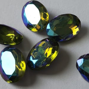 Pode incluir: Close-up de seis contas ovais iridescentes. As contas têm uma superfície facetada que reflete a luz amarela, verde e azul. As contas estão espalhadas sobre uma superfície cinza claro. Estas contas são adequadas para fazer joias.