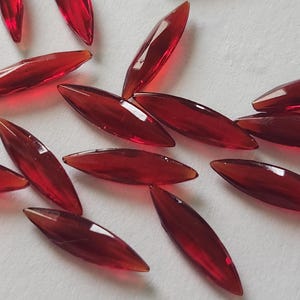 May include: A collection of translucent, elongated, red glass beads. Each bead has a pointed, leaf-like shape and a glossy finish. The beads are scattered on a white surface, creating a visually appealing arrangement.