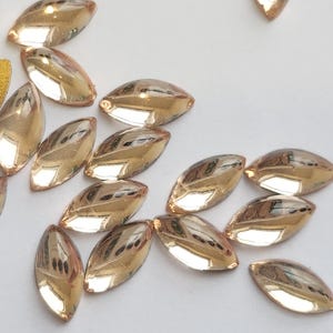 Puede incluir: Un grupo de 15 piedras de cristal de forma de lágrima de color dorado. Las piedras están esparcidas sobre una superficie blanca.