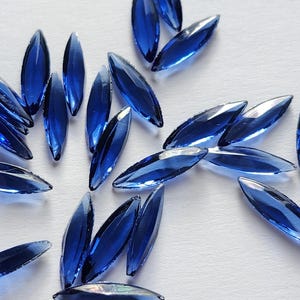 May include: A scattering of small, blue, glass, teardrop-shaped embellishments. The embellishments are scattered on a white surface.
