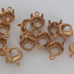 May include: A group of 12 gold-toned metal settings for cabochons. Each setting has a hexagonal shape with a small loop at the top for attaching to a chain or other jewelry piece.