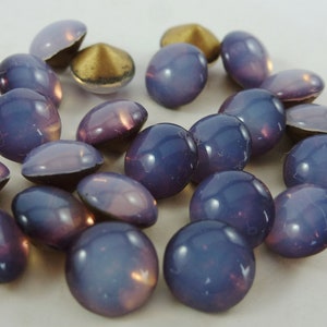 Op de afbeelding: Een verzameling iriserende, ovaalvormige kralen in blauwe en paarse tinten, met een metallic gouden achterkant. De kralen zijn verspreid, waarbij hun glanzende, reflecterende oppervlakken worden getoond. Deze kralen zijn geschikt voor het maken van sieraden en knutselprojecten.