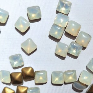 May include: Assortment of iridescent white and gold square-shaped rhinestones. The rhinestones are scattered on a white surface.