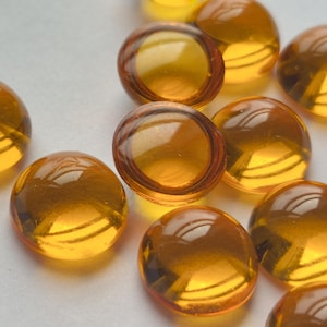 May include: A collection of small, round, translucent amber-coloured glass cabochons with a smooth, domed surface. These circular pieces feature a warm, golden-orange hue and a polished finish, reflecting light across their curved, semi-spherical tops.