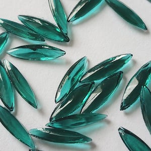May include: A collection of teal-colored, leaf-shaped glass beads scattered on a white surface. The beads are elongated with pointed ends and a glossy finish, reflecting light. These decorative beads are ideal for jewelry making and craft projects.