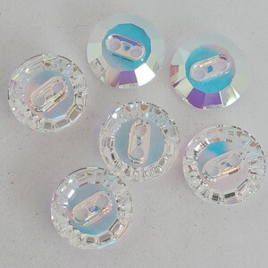 Puede incluir: Seis botones de cristal transparente iridiscente con un lazo de metal plateado en el centro. Los botones son redondos y tienen un diseño facetado.