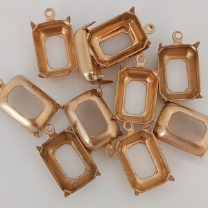 May include: A group of ten gold-toned metal settings for bezel settings. The settings are rectangular with a rounded edge and have a small loop at the top for attaching to a chain or other jewelry.