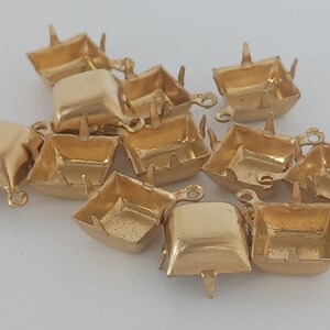 May include: A group of 12 gold-colored metal settings for cabochons. The settings are square shaped with four prongs on the top and a loop on the side.