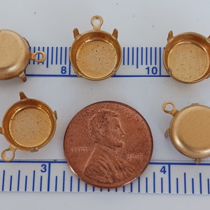 May include: A selection of small, gold-coloured metal jewellery findings. The findings include round bezels with loops and prongs, and a penny for size comparison. The items are arranged on a surface with a ruler.