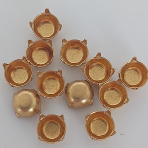 Puede incluir: Un grupo de 12 engastes metálicos de color dorado para diamantes de imitación u otros adornos. Los engastes tienen cuatro garras en la parte inferior y una parte superior redonda ligeramente cóncava.