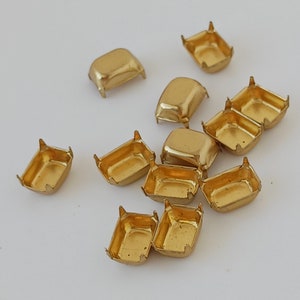 May include: A group of 11 gold-colored metal settings for rhinestones or other embellishments. The settings are rectangular with rounded corners and have four prongs on each side.