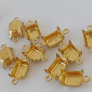 May include: A group of 10 gold-toned metal settings for cabochons. Each setting has a square shape with rounded corners and three prongs on each side. The settings are designed to hold a cabochon in place.