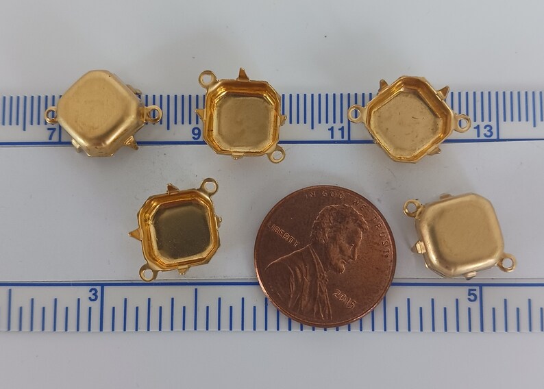 Może przedstawiać: Pięć złotych metalowych opraw do kaboszon&oacute;w, każda z trzema szponami i dwoma pętlami. Oprawy mają średnicę około 2,5 cm i są pokazane obok amerykańskiego centa dla skali.