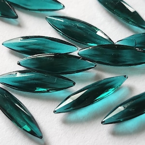 May include: A collection of teal-colored, teardrop-shaped glass beads. The beads are elongated with pointed ends and a slightly curved shape. They are scattered on a white surface, catching the light and reflecting a glossy sheen.