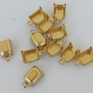 May include: A group of ten gold-toned metal settings for cabochon stones. The settings are in the shape of a rectangle with rounded corners and have a small loop on the top for attaching to a chain or other jewelry.