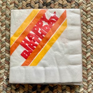 May include: White paper napkin with the words "Happy Birthday" printed in red and orange letters on a diagonal stripe pattern.