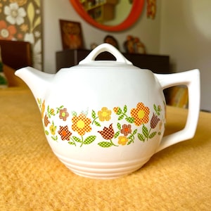 May include: White teapot with a floral pattern in shades of orange, yellow, brown, and green. The flowers are stylized and have a checkered pattern.