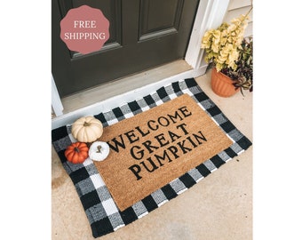 Puede incluir: Una alfombra de coco marrón con el texto "Welcome Great Pumpkin" en letras negras. La alfombra está sobre una alfombra a cuadros negros y blancos con tres pequeñas calabazas en la alfombra.